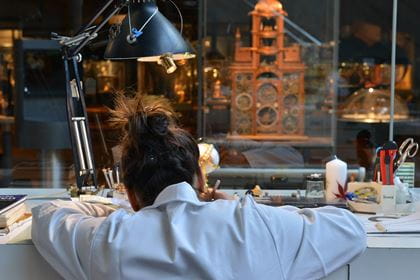 Eine Uhrmacherin ist von hinten zu sehen, wie sie am Arbeitsplatz sitzt und sich über etwas beugt. Vor ihrem Arbeitsplatz ist eine Glasscheibe, durch welche man sie bei der Arbeit beobachten kann.