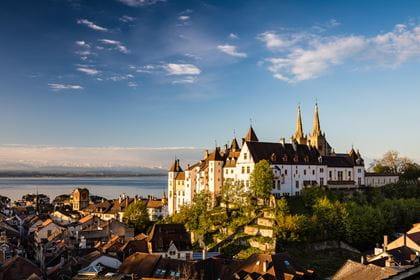 Das Schloss von Neuenburg mit dem See im Hintergrund. Rundherum befinden sich Häuser und Bäume. Der Himmel ist blau.