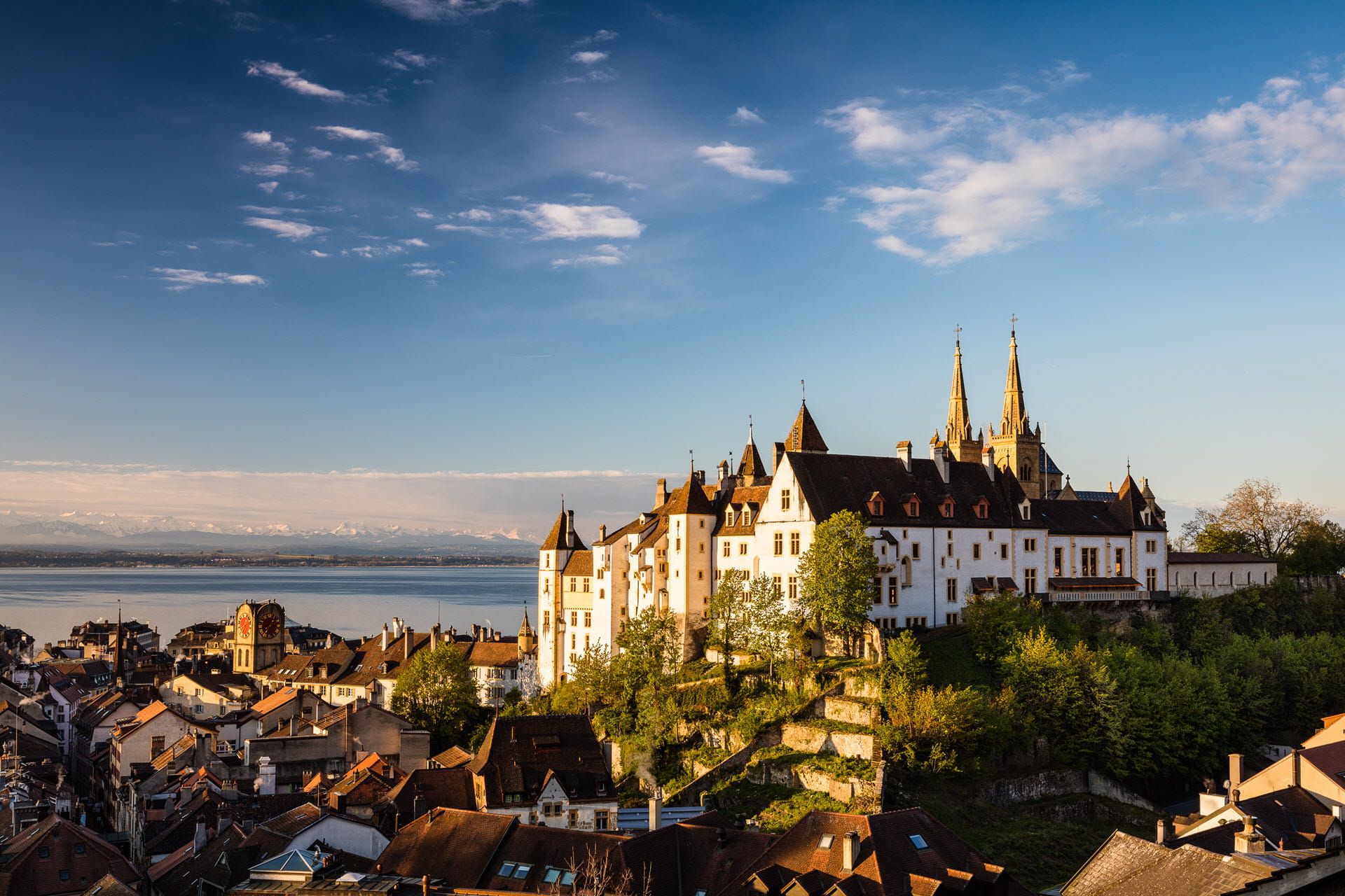 Le château de Neuchâtel avec le lac en arrière-plan. Tout autour, il y a des maisons et des arbres. Le ciel est bleu.