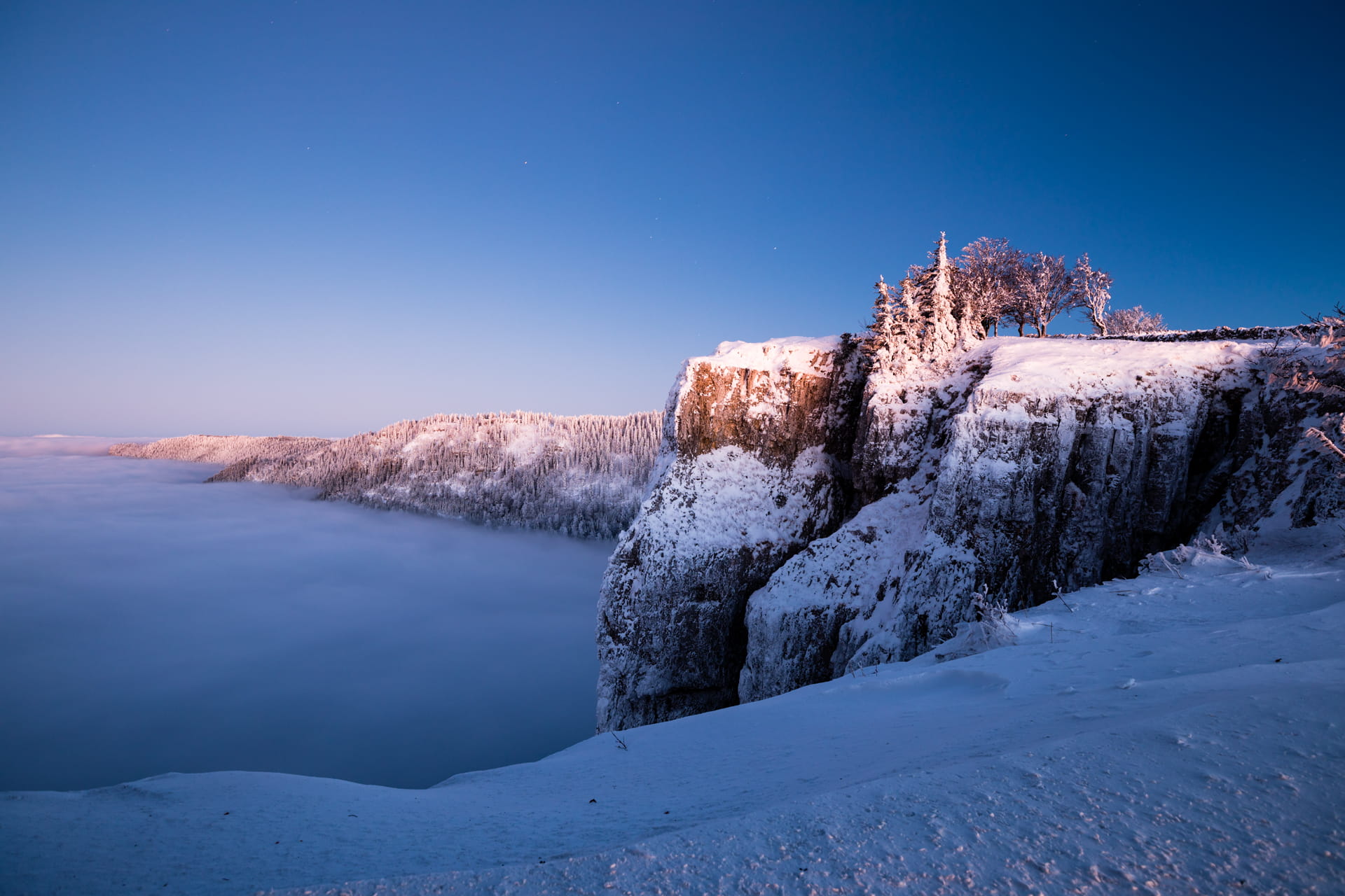 Man steht nahe am Abgrund des Creux du Van. Es ist Winter und der Boden sowie die schroffen Klippen rechts im Bild sind schneebedeckt. Man hat eine weite Aussicht: rechts zieht sich die Gebirgskette weiter durchs Bild, geradeaus blickt man auf ein Nebelmeer.