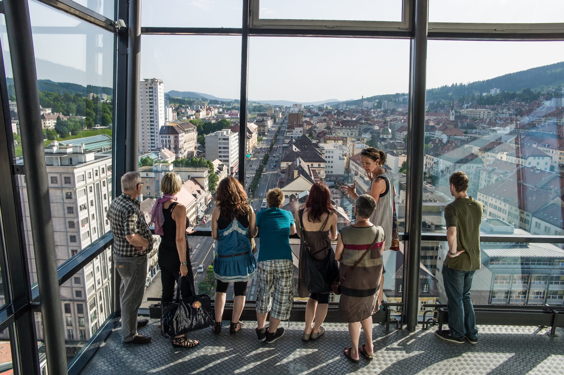 Un petit groupe se trouve sur le pont d'observation de la Tour de l'Espacité à La Chaux de Fonds. La ville est visible derrière les grandes fenêtres panoramiques. 