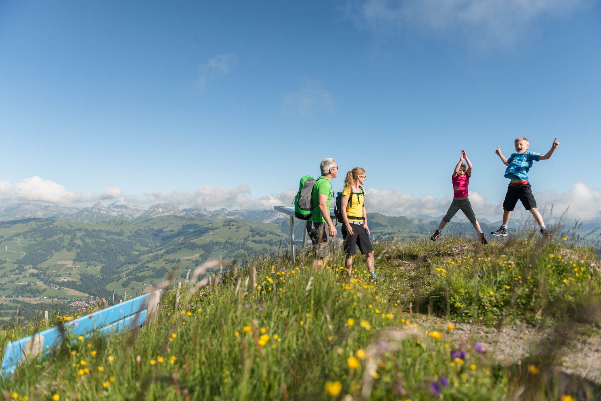 Die zwei Kinder der vierköpfigen Familie springen vor einem Bergpanorama in die Luft.