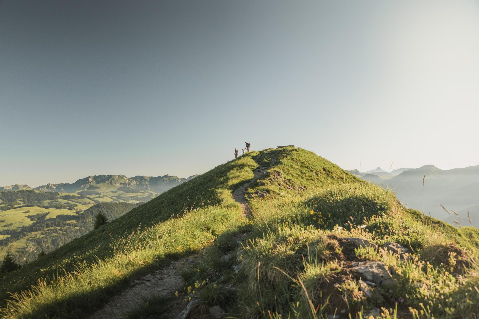Ein dünner Weg führt über den Gebirgskamm bis zu einem Aussichtspunkt mit Bänkli. Weit entfernt sieht man eine Familie die gerade den Abstieg in Angriff nimmt. © Melanie Uhkoetter