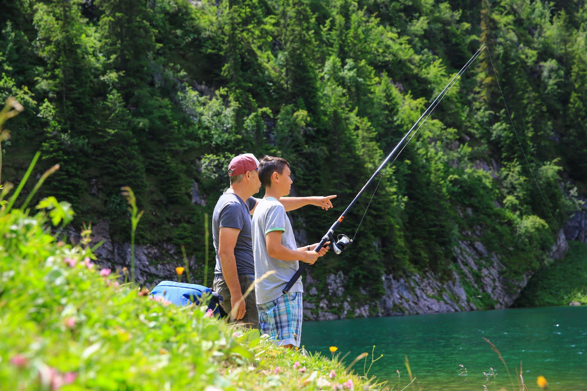 Vater und Sohn stehen am Seeufer und sind am Fischen. Das Foto wurde seitlich von rechts hinten aufgenommen. Der Vater trägt ein rot-weisses Basecap und zeigt mit dem Finger geradeaus. Der Sohn hält die Angelrute in den Händen.
